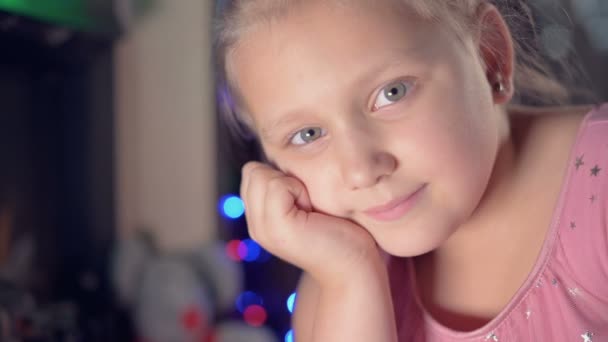 Uma menina em um vestido festivo contra o pano de fundo de luzes festivas e uma árvore de Natal sorridente olha para o borrão da câmera e movimento para se concentrar. O conceito de umas férias estragadas — Vídeo de Stock