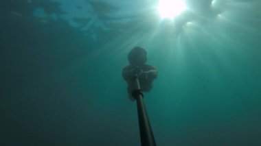 Maskeli ve şnorkeli olan genç bir sporcu suyun altında selfie çubuğuyla yüzüyor. Suyun içinde güneş parlıyor.