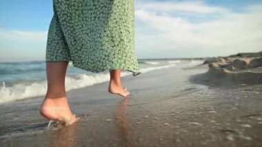 Sabahın erken saatlerinde sahilde çıplak ayakla yürüyen, ağır çekimde ince, çekici bacakları olan bir kadın. Altın plajda açık yeşil elbiseli bir turist