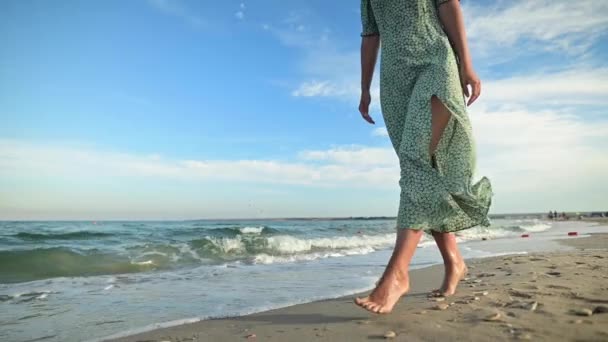 Atractiva mujer en cámara lenta caminando descalza a lo largo de la playa por la mañana temprano. Turista en un vestido verde claro en una playa dorada — Vídeo de stock