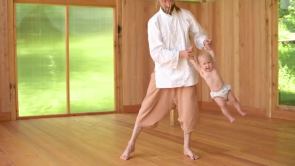 一位父亲，一位穿着浅色宽松衣服的瑜伽和气功修行者，和他一岁的女儿在练习场玩耍。带着吊儿郎当的体操表演 — 图库视频影像