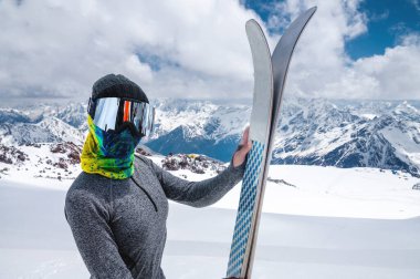 İnce Müslüman bir sporcunun portresi kapalı bir yüzle ellerinde kayaklarla Kafkasya 'nın yüksek dağlarının arka planına yaslanmış.