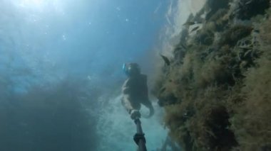 Maskeli genç adam batık bir teknenin yanında suyun altında yüzüyor. Selfie çubuğu aksiyon kamerası