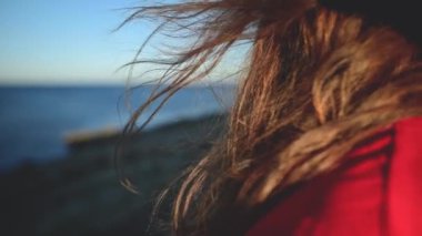 Deniz kenarındaki kırmızı elbiseli çekici beyaz kadın güneşe doğru bakıyor. Rüzgarda dalgalanan saçlar. Yavaş çekim alanının yüzeysel derinliği..