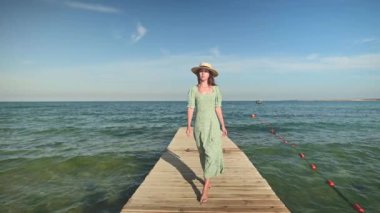 Gevşek yeşil elbiseli ve hasır şapkalı hoş bir beyaz kadın, güneşli bir günde mavi gökyüzü ve deniz dalgalarının zemininde, ahşap bir iskele ya da köprü boyunca yürüyor. Yavaş çekim