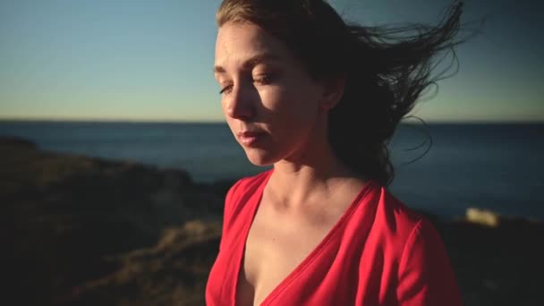 迷人的白种人年轻女人穿着红色的衣服在海边望着太阳。头发在风中飘扬近距离浅浅的场慢动作. — 图库视频影像
