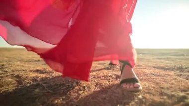 Yakın plan kadın sandaletler içinde ince bacaklar rüzgarda dalgalanan şeffaf kırmızı elbiseler doğadaki kuru çimenlere gider. Düşük açı ağır çekim. Parıltılı arka plan ışığı