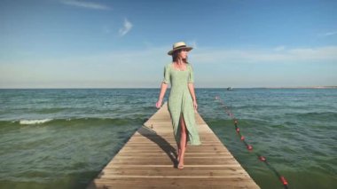 Gevşek yeşil elbiseli ve hasır şapkalı hoş bir beyaz kadın, güneşli bir günde mavi gökyüzü ve deniz dalgalarının zemininde, ahşap bir iskele ya da köprü boyunca yürüyor. Yavaş çekim