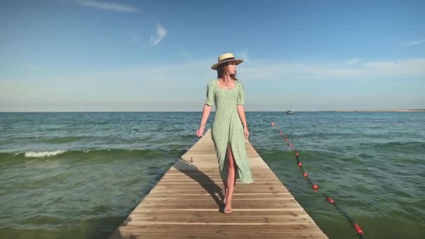 Una encantadora mujer caucásica joven con un vestido verde suelto y un sombrero de paja camina a lo largo de un muelle de madera o puente contra el telón de fondo del cielo azul y las olas del mar en un día soleado. Movimiento lento — Vídeo de stock