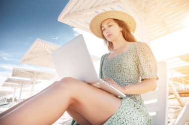 Yeşil elbiseli ve şapkalı çekici beyaz bir kadın, elinde dizüstü bilgisayarla bir şemsiyenin altında, okyanus yakınlarındaki kumlu bir sahilde dinleniyor. Serbest ve uzak çalışma