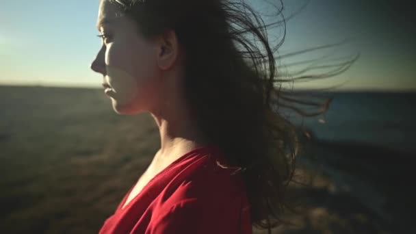 Attraente giovane donna caucasica in abito rosso in riva al mare guarda verso il sole. Capelli svolazzanti nel vento Close-up superficiale profondità di campo rallentatore. — Video Stock