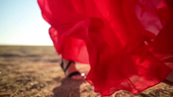 Close-up femminile Gambe slanciate in sandali in un abito rosso allentato trasparente che svolazza nel vento in onde va su erba secca in natura. Movimento lento ad angolo basso. Retroilluminazione con abbagliamento — Video Stock