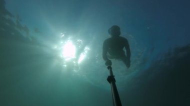 Maskeli ve şnorkeli olan genç bir sporcu suyun altında selfie çubuğuyla yüzüyor. Suyun içinde güneş parlıyor.