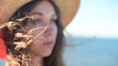 Kırmızı elbiseli ve deniz kıyısında hasır şapkalı çekici beyaz kadın ufka bakıyor. Rüzgarda dalgalanan saçlar. Yavaş çekim alanının yüzeysel derinliği..