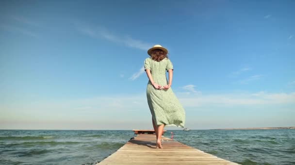 Una joven con un vestido verde de verano y un sombrero de paja en cámara lenta camina a lo largo de un muelle de madera por la mañana en el mar durante el viento. Caminar gracia belleza y feminidad — Vídeos de Stock