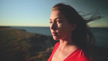 Deniz kenarındaki kırmızı elbiseli çekici beyaz kadın güneşe doğru bakıyor. Rüzgarda dalgalanan saçlar. Yavaş çekim alanının yüzeysel derinliği..
