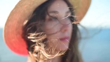 Kırmızı elbiseli ve deniz kıyısında hasır şapkalı çekici beyaz kadın ufka bakıyor. Rüzgarda dalgalanan saçlar. Yavaş çekim alanının yüzeysel derinliği..