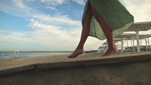 Vonzó karcsú lábai vannak egy lassú mozgású nőnek, aki mezítláb sétál a parton kora reggel. Turista egy világoszöld ruha egy arany strandon — Stock videók