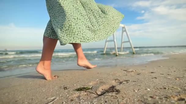 Atractivas piernas delgadas de una mujer en cámara lenta caminando descalza por la playa por la mañana temprano. Turista en un vestido verde claro en una playa dorada — Vídeo de stock
