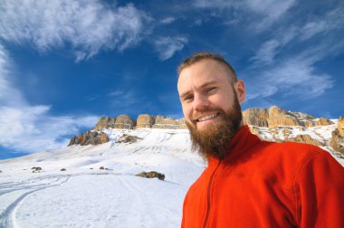 Sakallı neşeli genç adam dağların arka planında gülümsüyor.