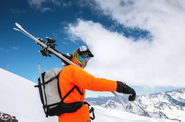 Genç adam omuzlarında kayaklarla dağların arka planında duruyor.