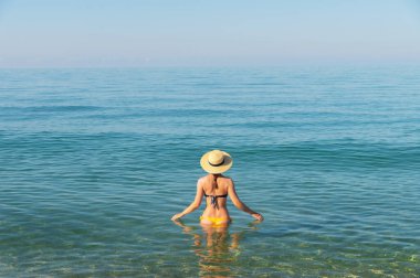 Güneşli bir günde, beline kadar deniz suyuna batmış, mayo giymiş, ince bir genç kadın.