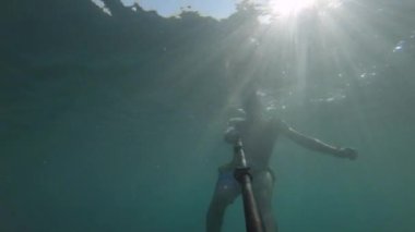Maskeli ve şnorkeli olan genç bir sporcu suyun altında selfie çubuğuyla yüzüyor. Suyun içinde güneş parlıyor.