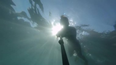 Maskeli ve şnorkeli olan genç bir sporcu suyun altında selfie çubuğuyla yüzüyor. Suyun içinde güneş parlıyor.