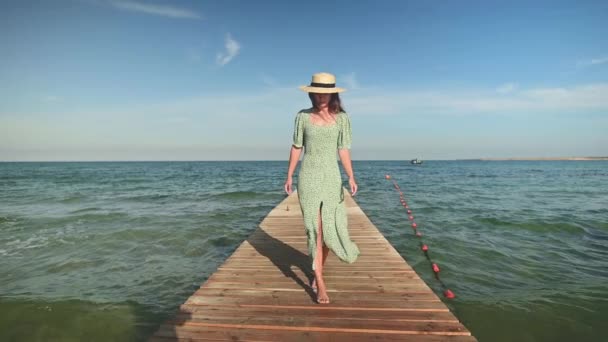 Μια νεαρή ελκυστική λευκή γυναίκα με πράσινο φόρεμα και ψάθινο καπέλο περπατά το καλοκαίρι σε μια ξύλινη προβλήτα στην παραλία. Χαλάρωση και χαλάρωση στις διακοπές και τα ταξίδια — Αρχείο Βίντεο