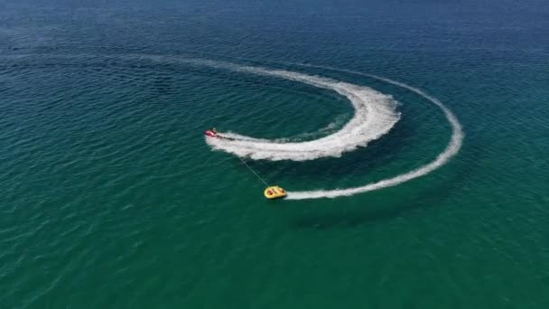 Vista aérea de tubos rebocados infláveis para rolar turistas em um tubo amarrado a jetski através do mar azul em um dia ensolarado — Vídeo de Stock