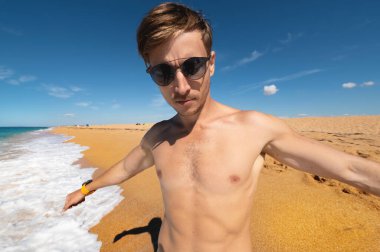 Güneş gözlüklü zayıf fizikli, çekici, beyaz, genç bir sporcu kollarını gösterişli bir jestle deniz kıyısında duruyor.