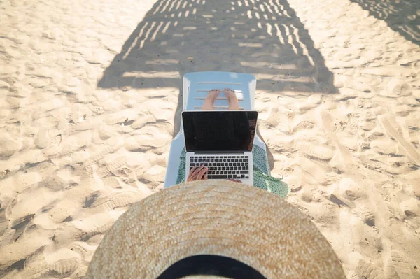 Верхний вид стройная молодая белая женщина в шляпе работает на его ноутбуке отдыхает на песчаном пляже в солнечный день. Тонкие ноги и шляпа на песчаном фоне — стоковое фото