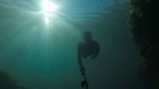 Młody sportowiec w masce i snorkel pływa z selfie kijem głęboko pod wodą. Słońce świeci przez wodę. — Wideo stockowe