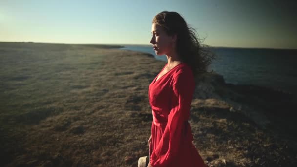 Attraente giovane donna caucasica in abito rosso in riva al mare guarda verso il sole. Capelli svolazzanti nel vento Close-up superficiale profondità di campo rallentatore. — Video Stock