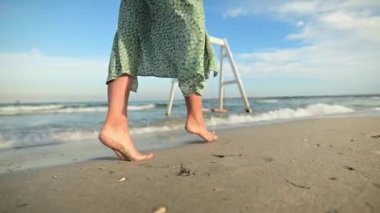 Yakından çıplak ayaklı yeşil elbiseli, çekici, beyaz tenli genç bir kadın sabahın erken saatlerinde deniz kenarında ıssız bir plaj boyunca geziniyor. Yavaş çekim.