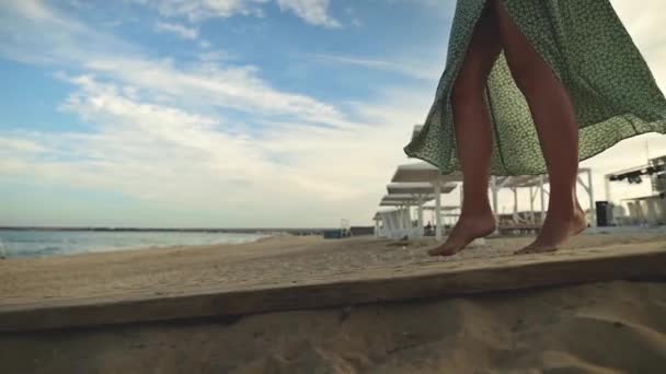 Uma jovem branca atraente e esbelta em um vestido verde solto passeia descalça ao longo de uma praia deserta perto do mar no início da manhã. Movimento lento — Vídeo de Stock