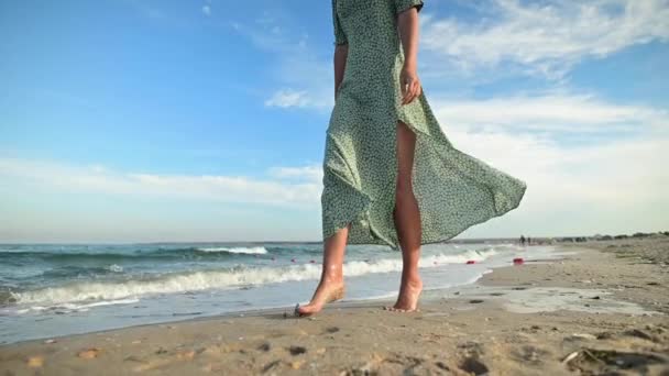 Uma jovem branca atraente e esbelta em um vestido verde solto passeia descalça ao longo de uma praia deserta perto do mar no início da manhã. Movimento lento — Vídeo de Stock