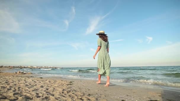 An attractive Caucasian slender young woman in a loose green dress strolls barefoot along a deserted beach near the sea in the early morning. Slow motion — Stock Video