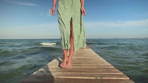 Primer plano vista baja joven atractiva esbelta mujer caucásica en un vestido verde y un sombrero de paja camina en el verano a lo largo de un muelle de madera en la orilla del mar. Relajación y relajación en vacaciones y viajes — Vídeos de Stock