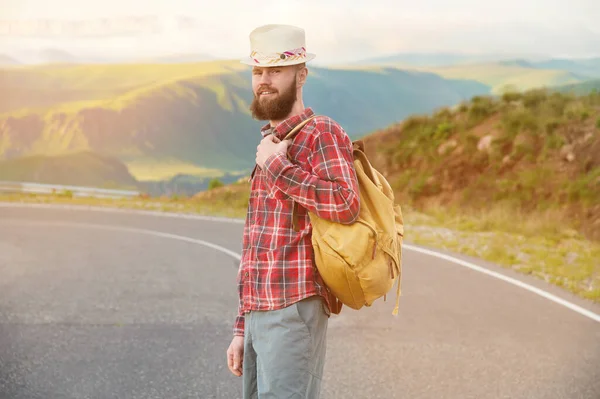 Homem barbudo em um chapéu fica na estrada contra o fundo das montanhas — Fotografia de Stock