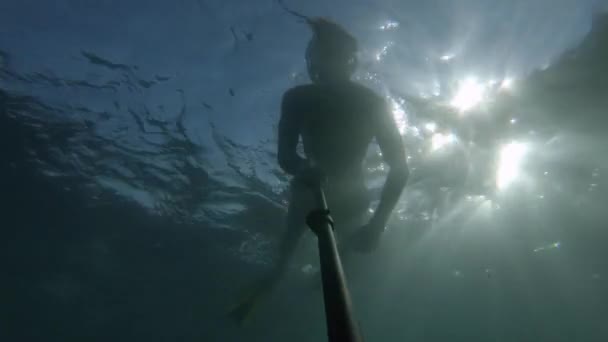 Un joven deportista con una máscara y un snorkel nada con un palo selfie profundo bajo el agua. Deslumbramiento del sol a través del agua — Vídeos de Stock