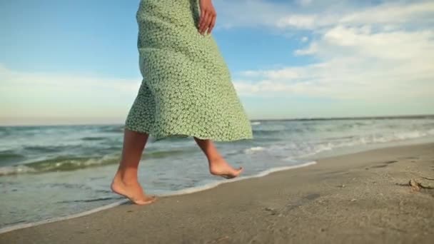 Pies desnudos de cerca Una atractiva joven blanca delgada en un vestido verde suelto pasea por una playa desierta cerca del mar por la mañana temprano. Movimiento lento. — Vídeos de Stock
