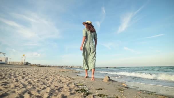 Atrakcyjna, biała, szczupła młoda kobieta w luźnej zielonej sukience spaceruje boso po bezludnej plaży nad morzem wczesnym rankiem. Zwolniony ruch — Wideo stockowe