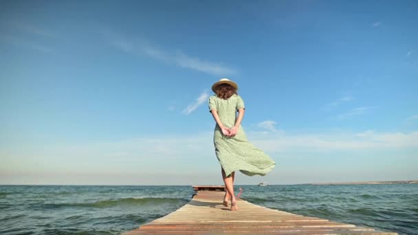 Молодая привлекательная стройная белая женщина в зеленом платье и соломенной шляпе ходит летом по деревянному пирсу на берегу моря. Отдых и отдых в отпуске и путешествиях — стоковое видео