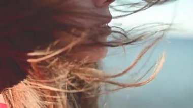 Rüzgarda yavaş çekimde dalgalanan Kafkasyalı genç bir kadının yakın çekimi. Yüzeyin derinliği. Kırmızı elbiseli ve şapkalı kız deniz kenarında yürüyor.