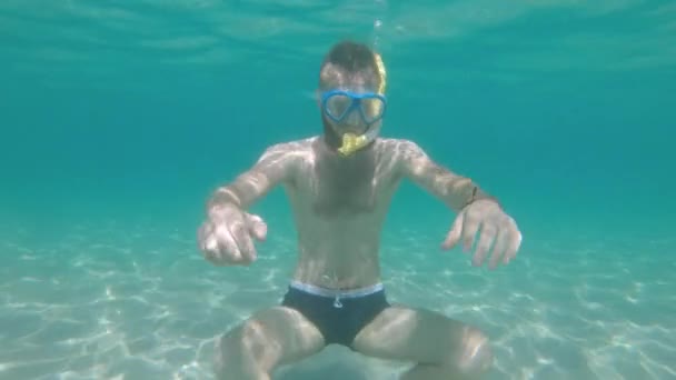 Undervattensbilder ung man med skägg i badbyxor under vattnet mask och snorkel försöker meditera på sandbotten av havet på en solig dag med bländning från vattenytan — Stockvideo