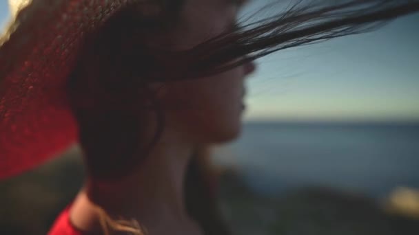 Primer plano de una joven mujer caucásica pelo revoloteando en cámara lenta en el viento. Profundidad de campo superficial. Chica en vestido rojo y sombrero soñador caminar por el mar — Vídeos de Stock