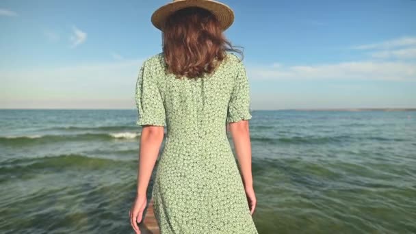 Close-up vista posteriore giovane attraente donna caucasica snella in un abito verde e un cappello di paglia passeggiate in estate lungo un molo di legno sulla riva del mare. Relax e relax in vacanza e in viaggio — Video Stock