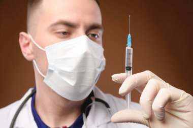 Kafkasyalı bir doktor elinde şırınga, elinde enjeksiyon ve açık bir ampul tutuyor. Diğer elinde de eldiven var. Aşılar