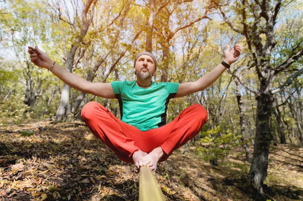 Um homem barbudo em equilíbrio de idade, sentado em uma tensa slackline na floresta de outono. Lazer ao ar livre — Fotografia de Stock